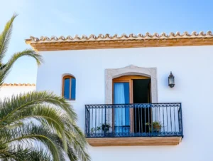 Balkon eines weißen mediterranen Hauses mit schmiedeeisernem Geländer und Pflanzen, daneben eine Laterne.