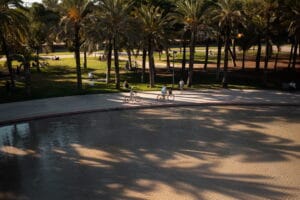 Zwei Personen fahren bei Sonnenlicht mit dem Fahrrad an einer Wasserfläche entlang, Menschen entspannen im Park