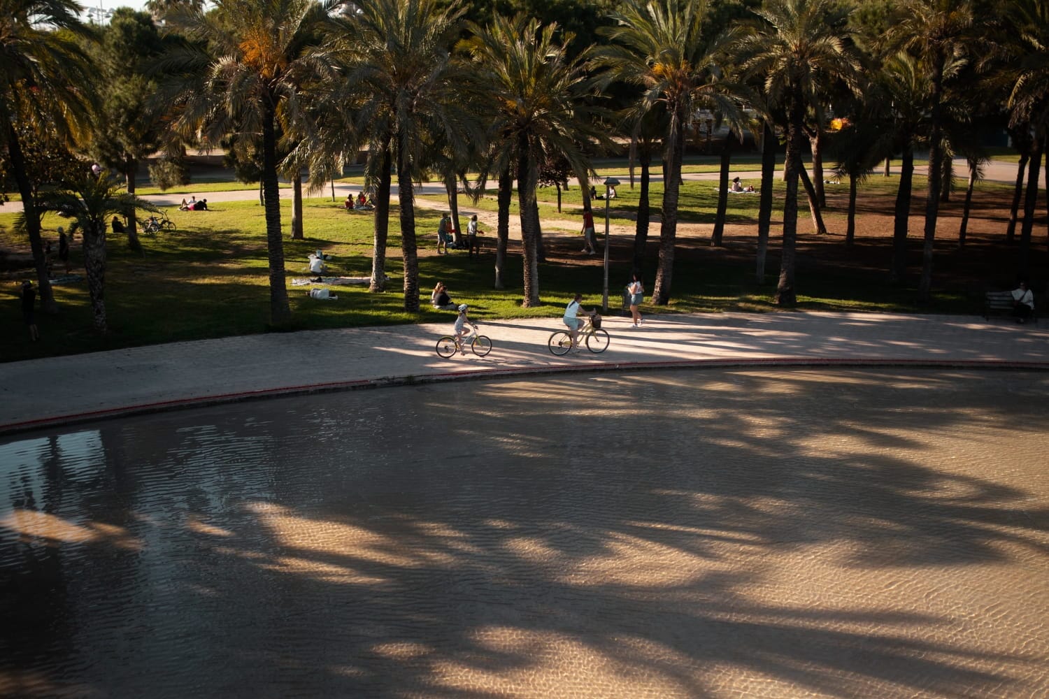 Zwei Personen fahren bei Sonnenlicht mit dem Fahrrad an einer Wasserfläche entlang, Menschen entspannen im Park