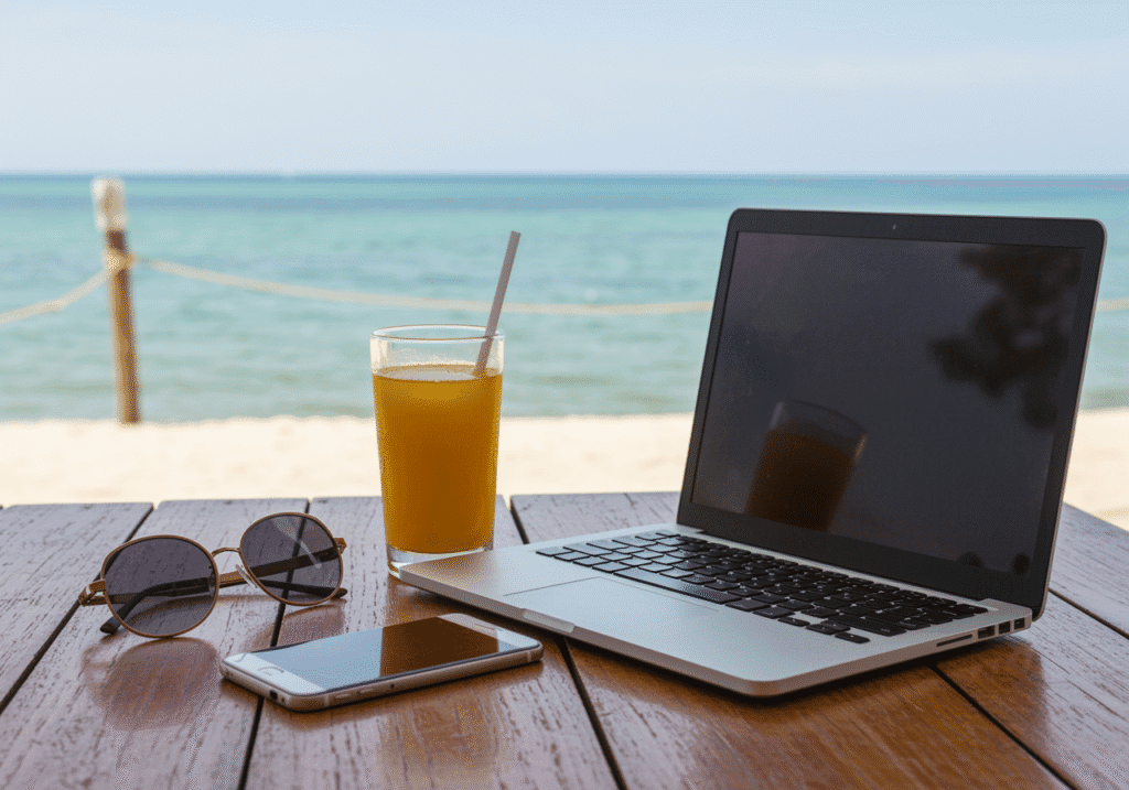 Laptop, Sonnenbrille, Smartphone und Orangensaft auf einem Holztisch am Strand mit Meerblick.