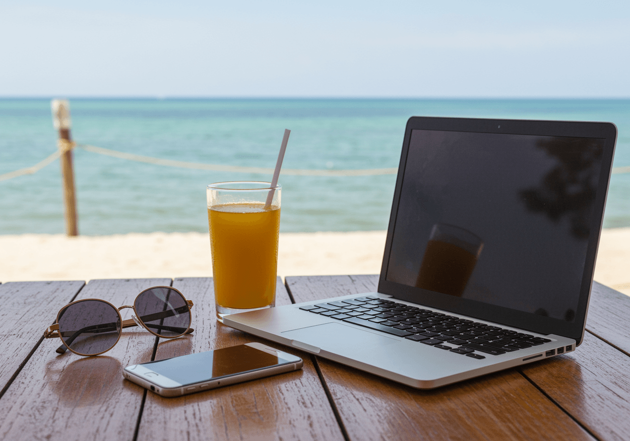 Laptop, Sonnenbrille, Smartphone und Orangensaft auf einem Holztisch am Strand mit Meerblick.